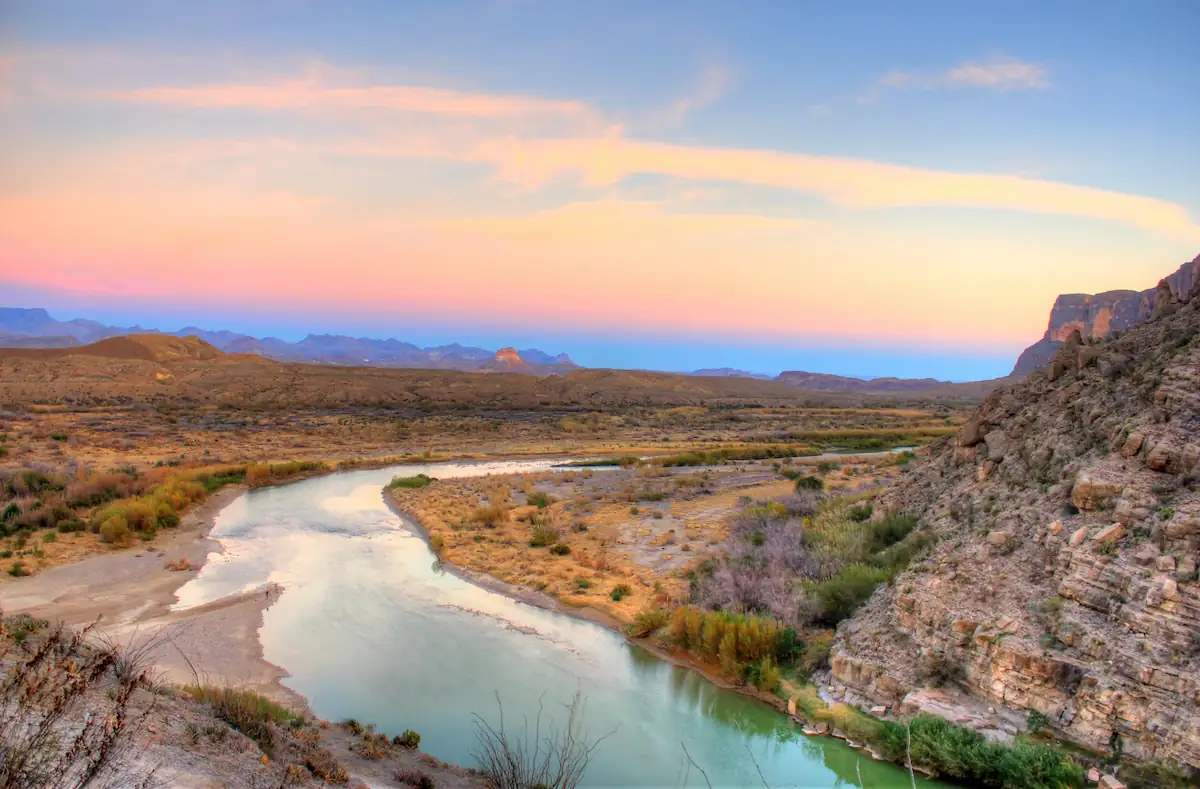 10 Affordable Activities for Kids in Texas 11 A serene landscape featuring a river winding through a rugged, arid terrain at sunset. The sky displays a gradient of colors from blue to pink, casting a warm glow over the distant hills and rocky cliffs lining the riverbanks. Sparse vegetation dots the scene.