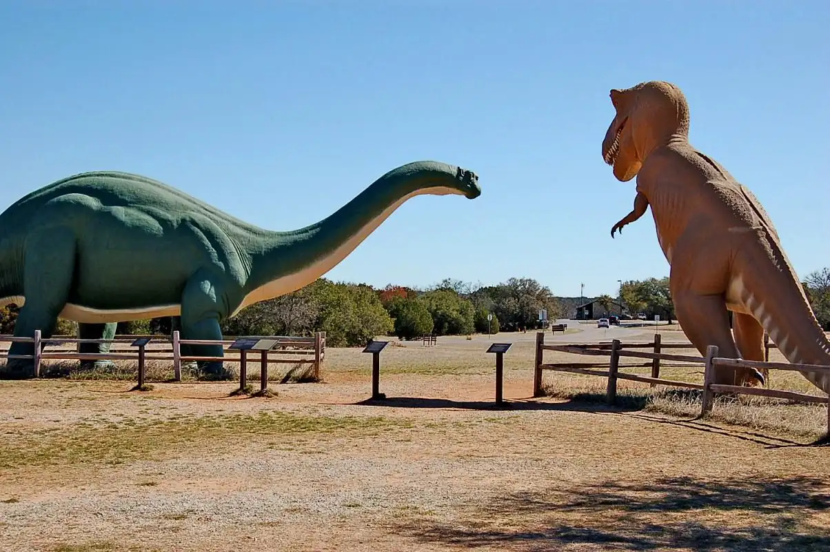 10 Affordable Activities for Kids in Texas 4 Two large dinosaur sculptures stand in a fenced outdoor area. The green one on the left is a long-necked sauropod, and the tan one on the right is a bipedal theropod. Information plaques are in front of each statue, and trees and buildings are visible in the background.