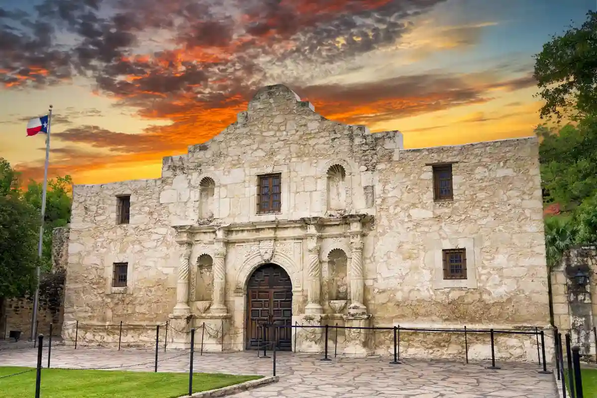 10 Affordable Activities for Kids in Texas 7 A historic stone mission stands under a vibrant sunset sky with wisps of orange, red, and yellow clouds. The mission features an arched entrance, wooden doors, and small windows. A flagpole with a colorful flag is visible on the left side of the image.