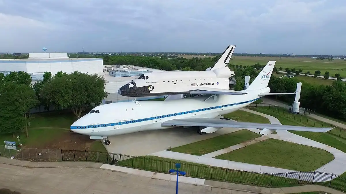 10 Affordable Activities for Kids in Texas 2 Aerial view of a display at NASA Johnson Space Center, featuring a large airplane with a space shuttle mounted on top. The structure is set against a backdrop of buildings and greenery under a cloudy sky.