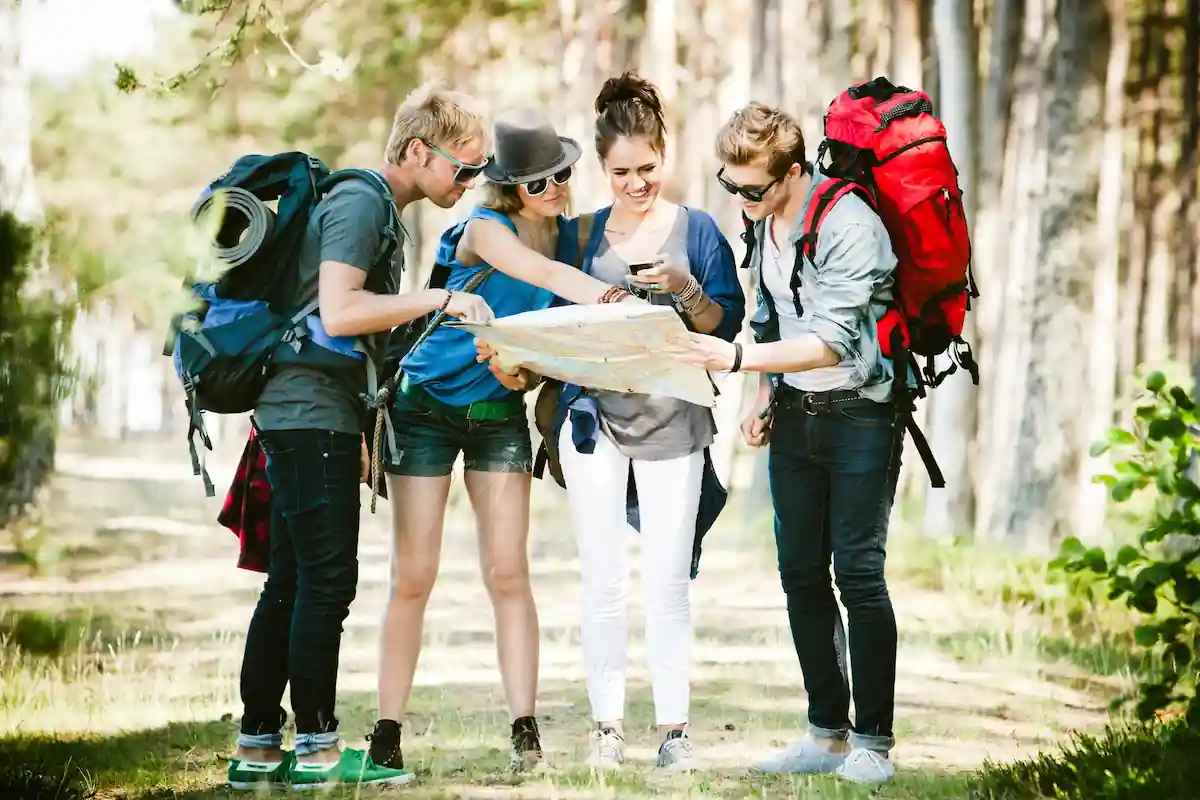 Can I Travel Internationally With A Misdemeanor: Yes or No? 2 Four young adults outdoors with backpacks, standing closely together and looking at a map, surrounded by trees. One person holds a paper map, and another appears to be checking a phone, all smiling and dressed in casual hiking gear.