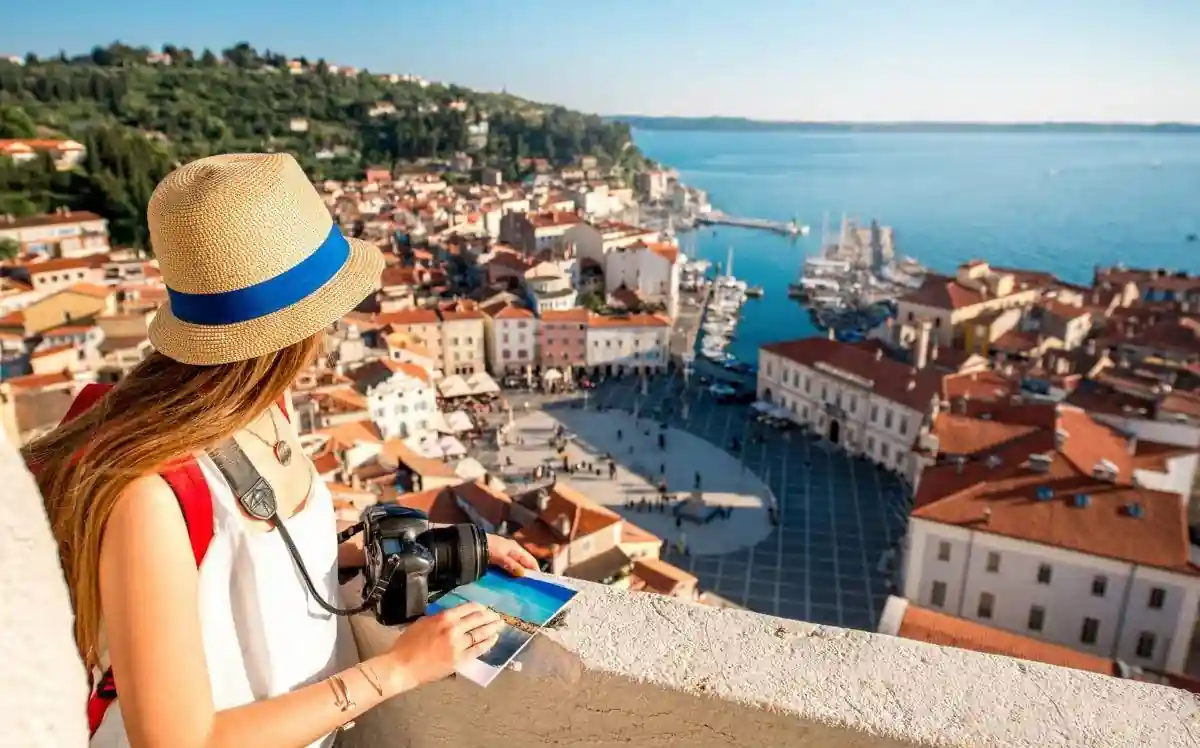 Can I Travel Internationally With A Misdemeanor: Yes or No? 4 A woman in a hat and tank top stands on a balcony, holding a camera and looking at a coastal town below. The town has red-tiled roofs, a marina filled with boats, and is surrounded by greenery with a blue sea and sky in the background.