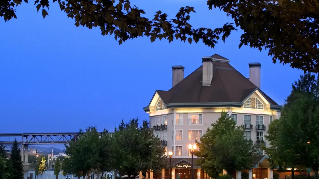 Where to Stay in Portland? 12 Top Rated Places in 2024 6 A photo of the Kimpton RiverPlace Hotel in Portland, Oregon. The hotel is a three-story building with a distinctive roofline and is illuminated against a twilight sky. Trees and a bridge are visible in the background.