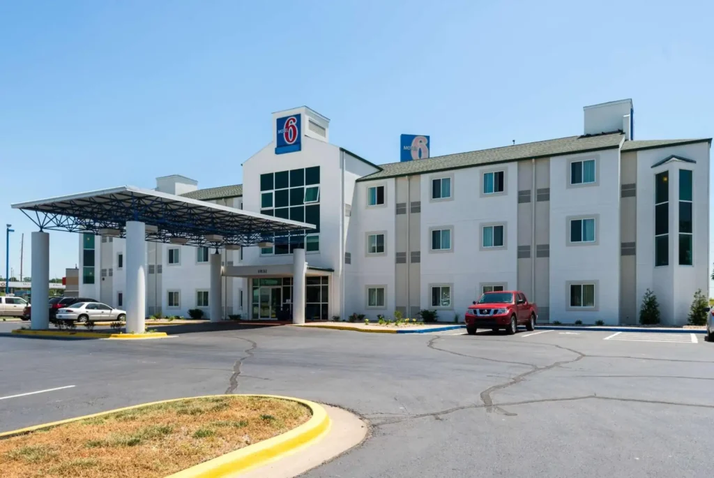 Where to Stay in Portland? 12 Top Rated Places in 2024 4 A white, two-story Motel 6 building with a blue awning over the entrance. The building has multiple windows and a large, rectangular sign featuring the Motel 6 logo and a cartoon dog. Cars are parked in the motel's parking lot. The motel is located in a suburban area with visible road signs and power lines.
