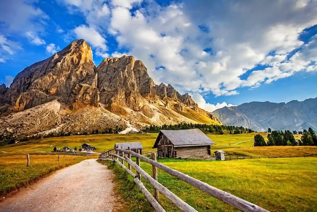 Can I Travel Internationally With A Misdemeanor: Yes or No? 6 A scenic mountain landscape featuring a dirt path lined with a wooden fence leading to rustic cabins. The backdrop showcases rugged mountains under a partly cloudy sky, with lush green fields in the foreground.