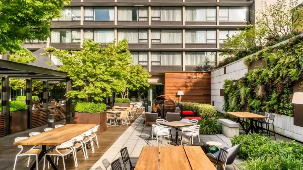 Where to Stay in Portland? 12 Top Rated Places in 2024 11 A photo of the Hotel Zags Portland's outdoor patio. The patio features a large, rectangular wooden table with chairs, a fire pit, and lush greenery, including trees, shrubs, and a vertical garden. There is also a covered seating area with a glass roof. The hotel building is visible in the background.