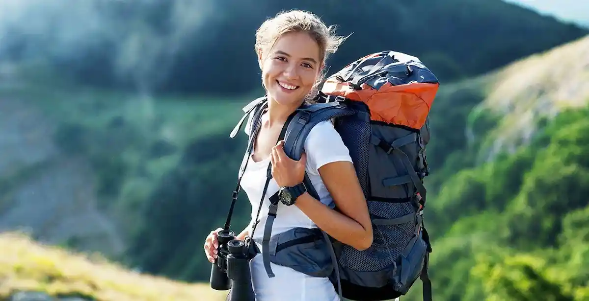 Can I Travel Internationally With A Misdemeanor: Yes or No? 9 A smiling person with blonde hair stands outdoors, wearing a large gray and orange hiking backpack and holding binoculars. The background features green hills under a clear sky, suggesting a scenic hike in nature.