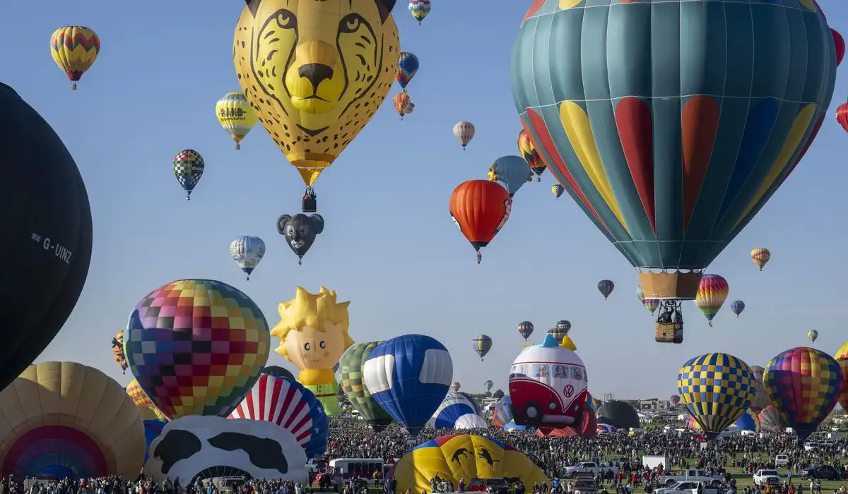 What to Expect at the Albuquerque Balloon Festival 2025 1 A vibrant scene of numerous hot air balloons filling the sky, featuring a variety of patterns and designs, including a cheetah face, a cow print, and colorful geometric shapes. Spectators gather on the ground in a green field under a clear blue sky.