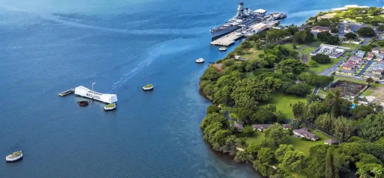 Aerial view of Pearl Harbor, showing the USS Arizona Memorial in the water and the Battleship Missouri docked nearby, surrounded by lush green trees and buildings along the shoreline.
