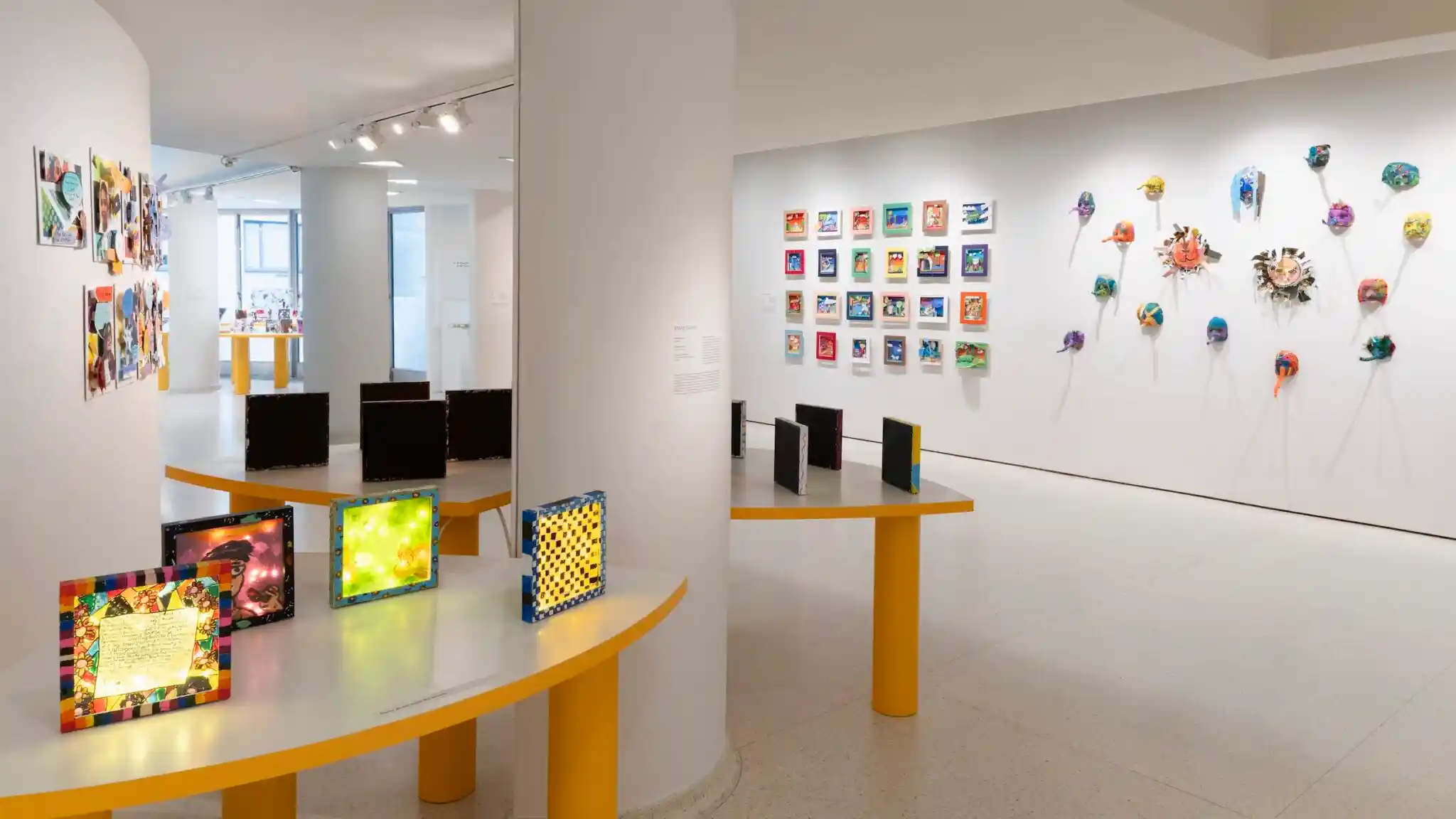 How to Visit The Gugg Like a Pro: Tips for Travelers 1 A brightly lit art gallery displays colorful framed artworks on white walls, vibrant 3D masks, and illuminated panels on yellow tables, creating a lively and modern exhibition space.