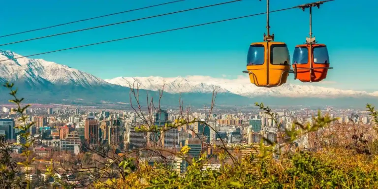 Two yellow cable cars travel above a city with modern buildings, lush greenery, and snow-capped mountains in the background under a clear blue sky.