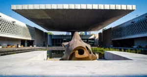 A large stone sculpture sits in a shallow water feature under a wide, flat, modern canopy at the National Museum of Anthropology in Mexico City, with white buildings surrounding the open courtyard.