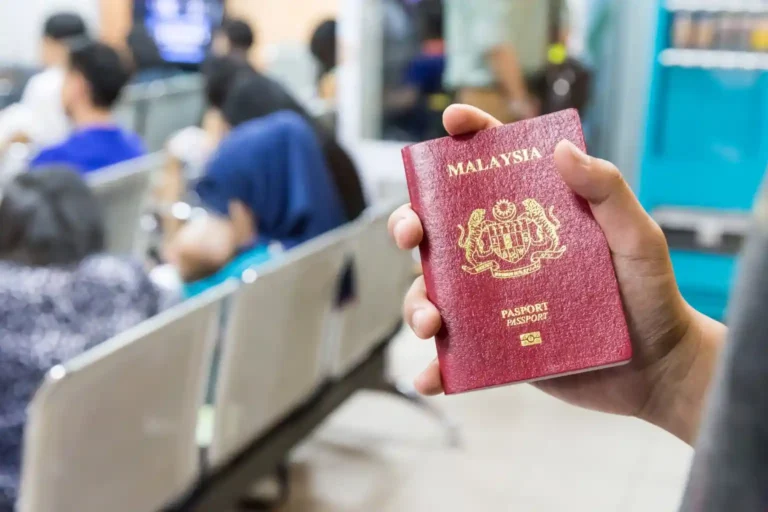 A person holds a Malaysian passport in their hand while sitting in a waiting area with other people seated in the background.