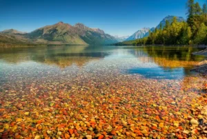 Clear lake with colorful pebbles visible underwater in the foreground, surrounded by green trees and distant mountains under a bright blue sky.