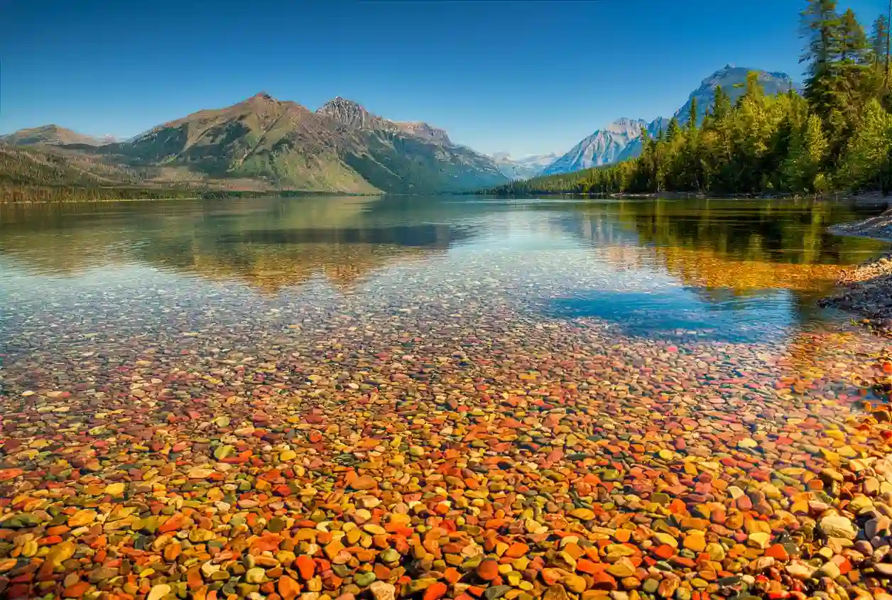 Clear lake with colorful pebbles visible underwater in the foreground, surrounded by green trees and distant mountains under a bright blue sky.