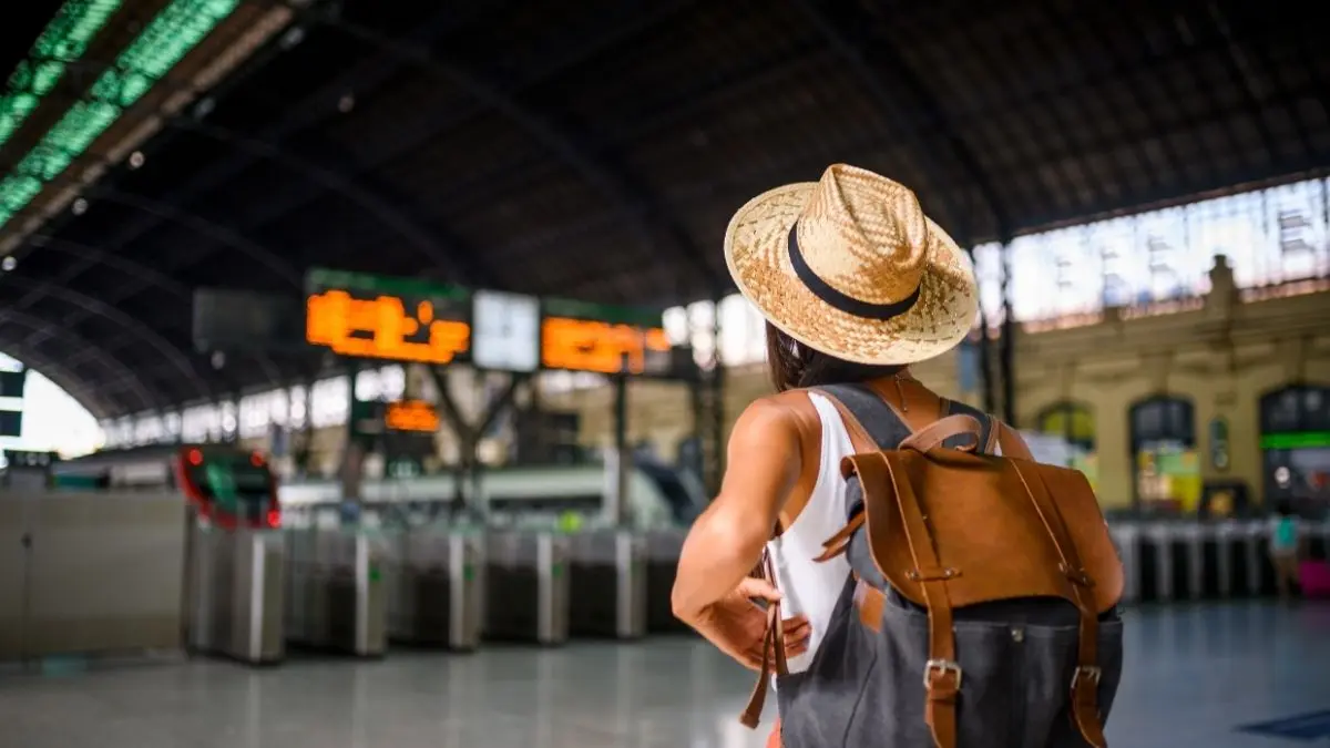 A traveler wearing a straw hat and backpack stands inside a large train station, facing electronic departure boards and turnstiles, with sunlight streaming through the windows above.