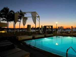 A serene poolside scene at dusk with glowing pool lights, empty lounge chairs, palm trees, and a sunset skyline over the water in the background.