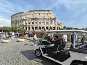 A white golf cart with people inside drives past a group of tourists near the Colosseum in Rome, Italy, on a sunny day with a partly cloudy sky.