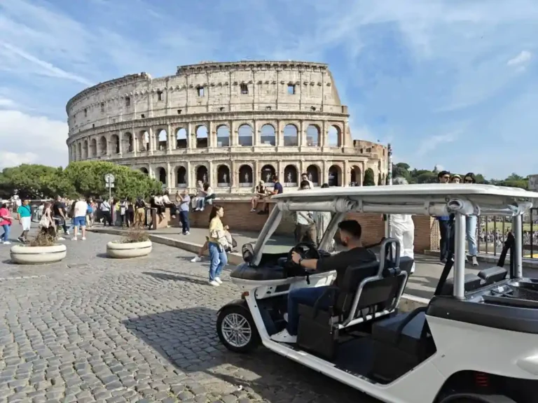 A white golf cart with people inside drives past a group of tourists near the Colosseum in Rome, Italy, on a sunny day with a partly cloudy sky.