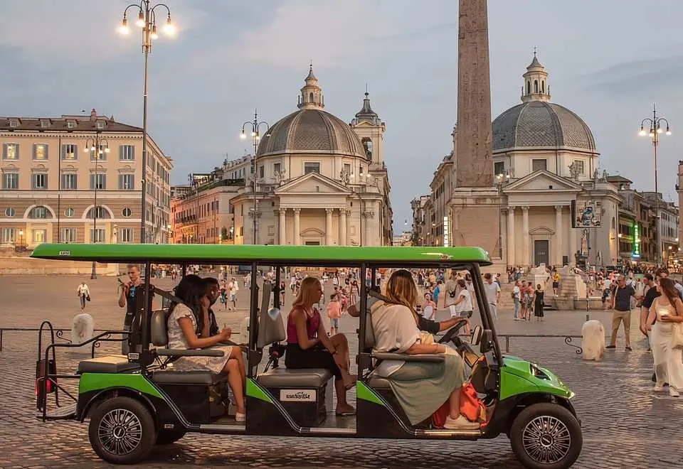 Rome by Golf Cart: The Easiest Way to See the Eternal City's Greatest Destinations 1 Rome by Golf Cart . A green electric tour cart with five people drives through a lively city square with historic domed buildings, an obelisk, and many pedestrians in the background during the evening.